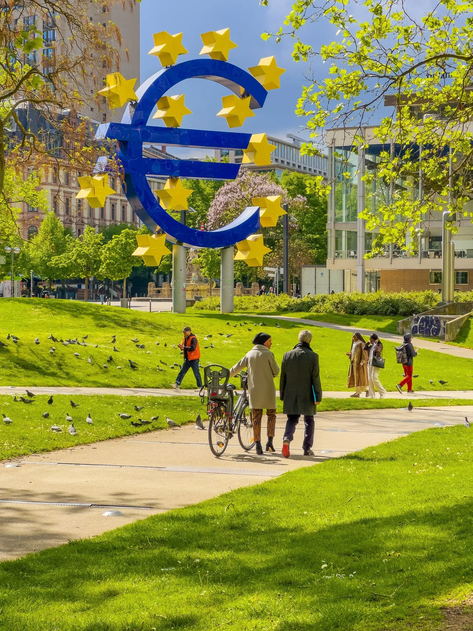 La escultura del euro en Fráncfort, un gran símbolo del euro azul rodeado de estrellas amarillas, con gente paseando por el parque alrededor